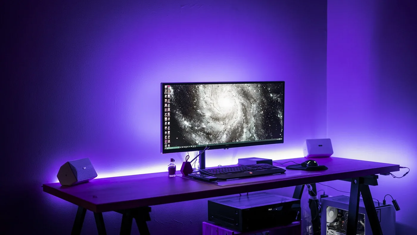 Un écran d'ordinateur ultra-large posé sur un bureau en bois, éclairé par un bandeau lumineux violet